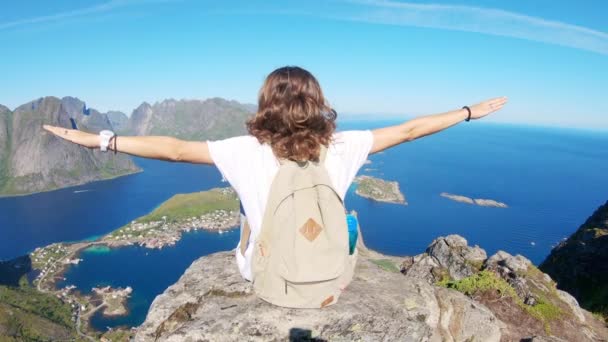 De jonge vrouw zit aan de rand van een klif met een bovenaanzicht naar de Reine — Stockvideo