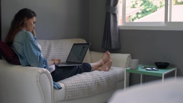 Uma jovem mulher com um laptop sentado no sofá na sala de estar — Vídeo de Stock