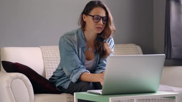 Een jonge vrouw met een laptop zittend op een bank en houdt een online consult. — Stockvideo