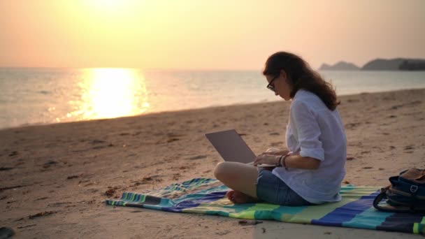 En ung kvinnlig författare sitter på stranden med sin laptop och skriver en text. — Stockvideo