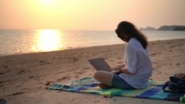 Uma jovem escritora sentada na praia com seu laptop e digitando um texto . — Vídeo de Stock