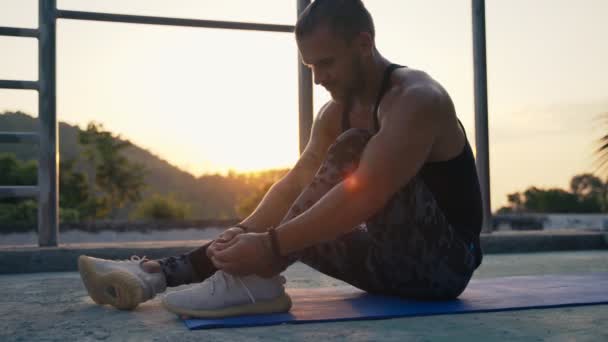 Атлетик сидит на спортивном коврике и завязывает шнурки. — стоковое видео