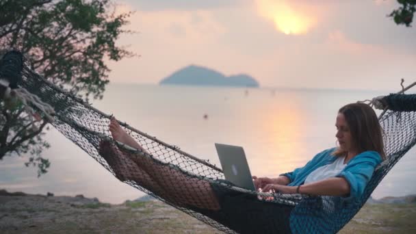 Молодая женщина-фрилансер лежит в гамаке с ноутбуком на тропическом пляже — стоковое видео