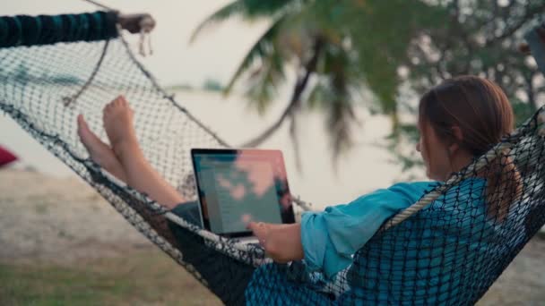 Молода жінка фрілансер лежить в гамаку з ноутбуком на тропічному пляжі — стокове відео