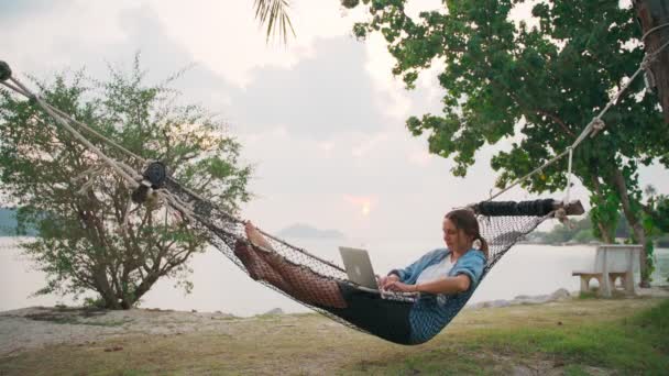 Mujer joven freelancer acostada en una hamaca con un portátil en la playa tropical — Vídeos de Stock