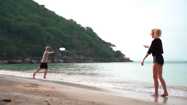 Två unga kvinnor kastar en frisbee på den tropiska stranden en solig dag. — Stockvideo