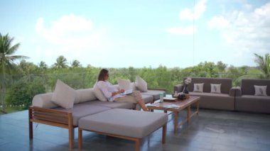 Dizüstü bilgisayarını kullanan genç bir kadın manzaralı rahat bir koltukta oturuyor.