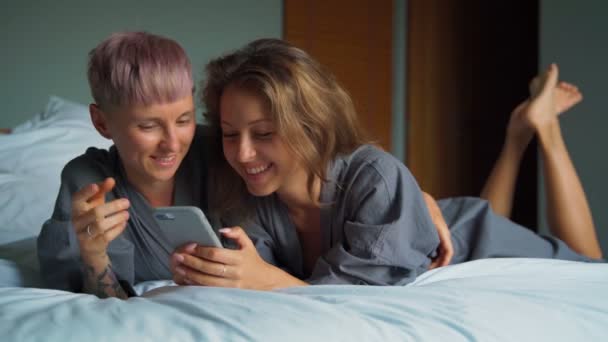 Fröhliches junges lesbisches Paar lacht, während es seine Smartphones im Bett benutzt — Stockvideo