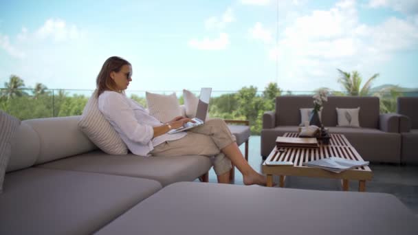 Jonge vrouw met behulp van haar laptop terwijl zitten op een comfortabele bank met een uitzicht — Stockvideo