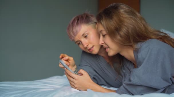 Fröhliches junges lesbisches Paar lacht, während es seine Smartphones im Bett benutzt — Stockvideo