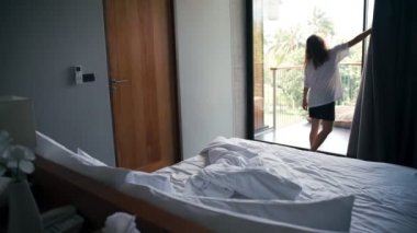 A young caucasian woman opens blackout curtains and goes onto a balcony