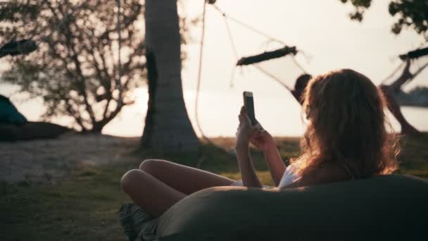 Young caucasian woman using her smartphone while lying on a big pillow — Αρχείο Βίντεο