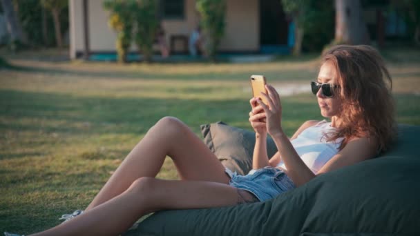 Молодая белая женщина использует свой смартфон, лежа на большой подушке — стоковое видео