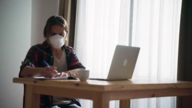 Maskeli genç bir kadın laptopunu kullanarak evden çalışıyor.