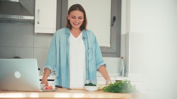 Молодая женщина готовит на кухне, разговаривая с друзьями через интернет . — стоковое видео