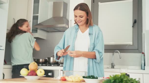 Két fiatal csinos nő énekel és táncol főzés közben a világos konyhában.. — Stock videók