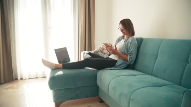 Jonge vrouw die thuis werkt met laptop en smartphone in de woonkamer. — Stockvideo