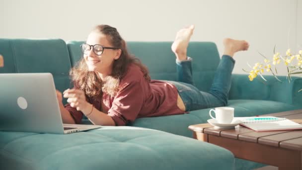 Eine junge, fröhliche Frau liegt mit einem Laptop auf einem blauen Sofa im Wohnzimmer — Stockvideo