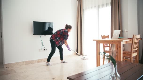 Μια ελκυστική νεαρή γυναίκα καθαρίζει το σαλόνι σφουγγαρίζοντας το πάτωμα. — Αρχείο Βίντεο