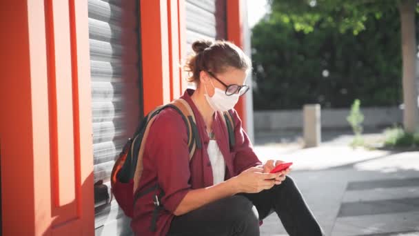 Koruyucu tıbbi maskeli ve gözlüklü genç bir kadın akıllı telefonunu kullanıyor. — Stok video