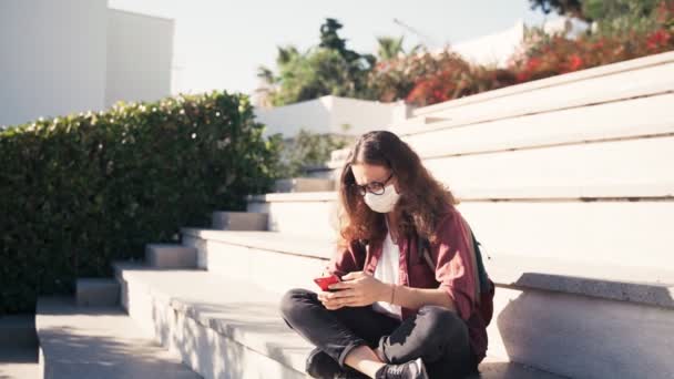 Una joven con una máscara médica y gafas usando su teléfono inteligente al aire libre — Vídeo de stock