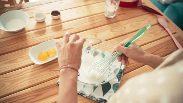 Una mujer batiendo huevos blancos con un batidor en un tazón de vidrio y añadiendo azúcar — Vídeo de stock