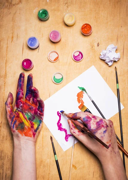 Hände des Künstlers in heller Farbe mit Pinsel auf Holztisch. Frau zeichnet auf einem Blatt Papier — Stockfoto