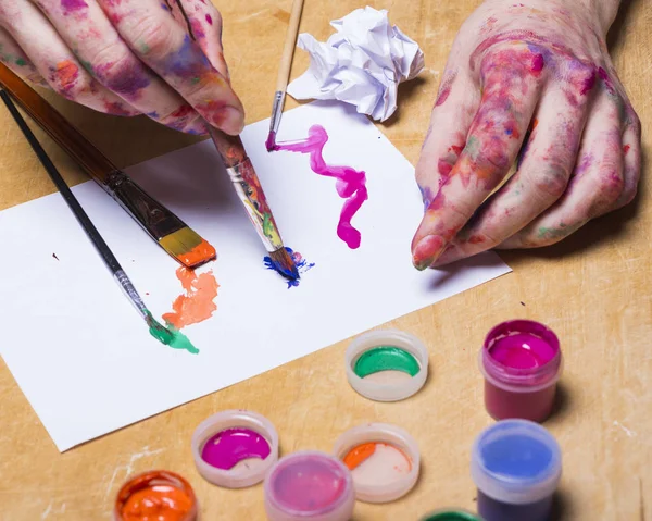 Manos del artista en pintura de color brillante con pincel sobre mesa de madera. Mujer dibuja en una hoja de papel — Foto de Stock
