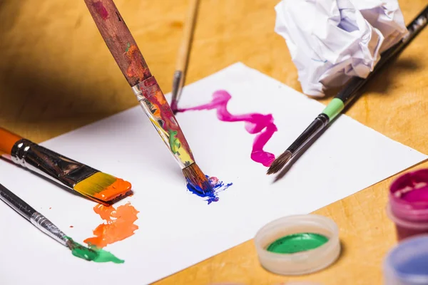 Manos del artista en pintura de color brillante con pincel sobre mesa de madera. Mujer dibuja en una hoja de papel — Foto de Stock