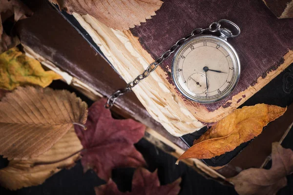 Reloj decadente en los viejos libros sabios de mala calidad con hojas amarillas secas de otoño sobre el fondo de madera oscura — Foto de Stock
