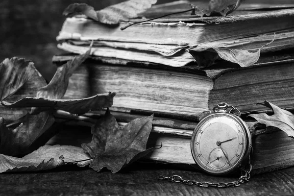 Schwarz-Weiß-Fotografie. verfallende Uhr auf den alten schäbigen klugen Büchern mit herbsttrockenem Laub auf dem dunklen Holzgrund — Stockfoto