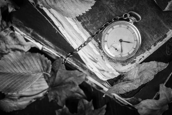 Черно-белая фотография. Разлагающиеся часы на старых потрепанных мудрых книгах с осенними сухими листьями на темном деревянном фоне — стоковое фото