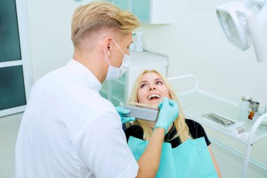 Dentist tries to choose color of the teeth and client clipart