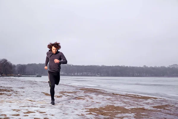 Young curly man running along the shore of a frozen lake in  win