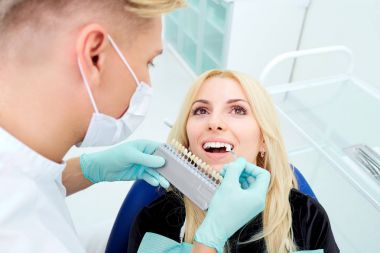 Dentist tries to choose the color of teeth and client in office clipart