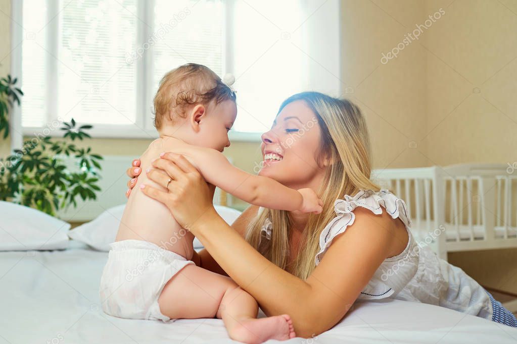 Mother and baby in a diaper play hugging on a bed indoors. 