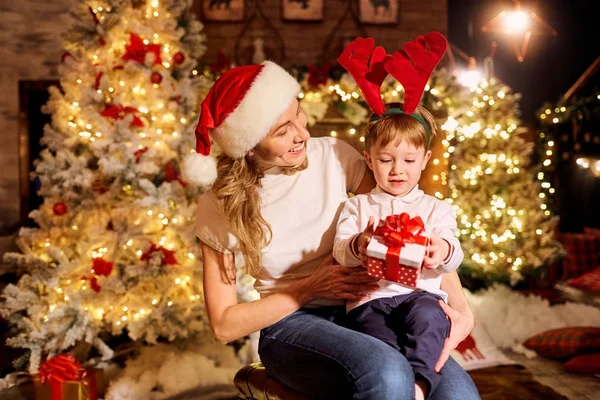 Mamá le da un regalo en una caja a su hijo en una habitación el día de Navidad . — Foto de Stock