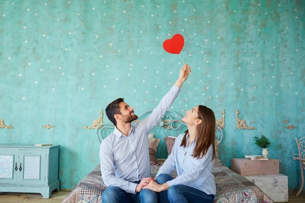 Um casal com corações nas mãos sentado na cama rindo — Fotografia de Stock