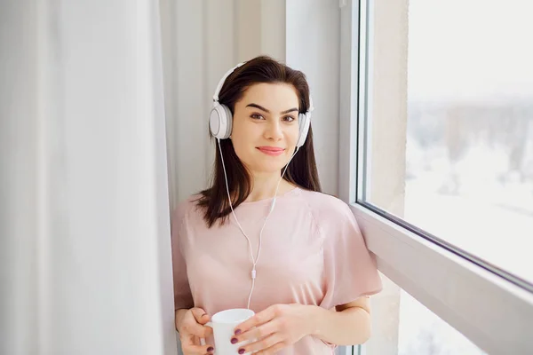 Una joven en auriculares con una taza en las manos junto a la ventana — Foto de Stock