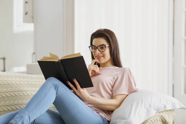 Une fille en lunettes lit le livre assis sur le canapé dans la chambre . — Photo