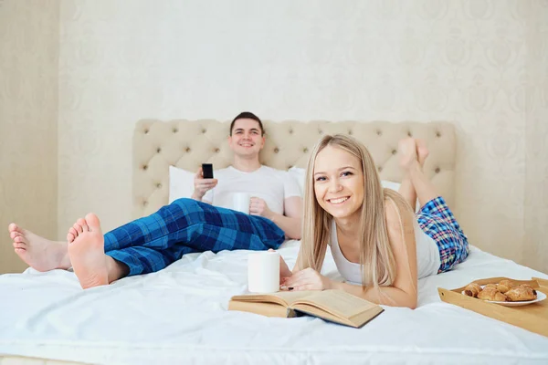 Para w łóżku jest o śniadanie, oglądanie telewizji, czytanie książki — Zdjęcie stockowe