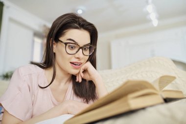 Kitap okurken kanepede yatan gözlüklü bir kız.