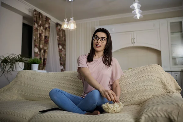 A brunette girl wearing glasses with popcorn watching TV  in the — Stock Photo, Image