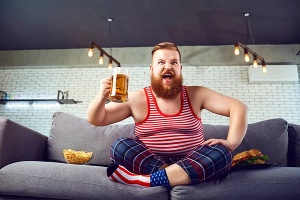 Spessore uomo divertente mangiare un hamburger seduto sul divano contro il — Foto Stock