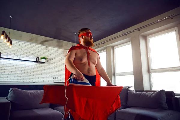 Grueso hombre divertido en un disfraz de superhéroe planchar cosas en casa . — Foto de Stock