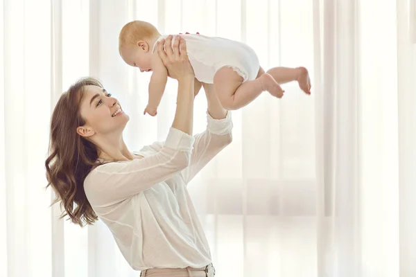 Madre con un bebé en sus brazos juega contra la ventana . —  Fotos de Stock