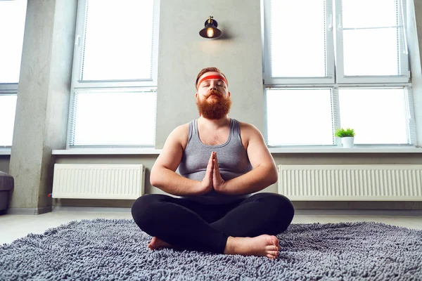 A funny fat bearded man in sports clothes does yoga in the room.
