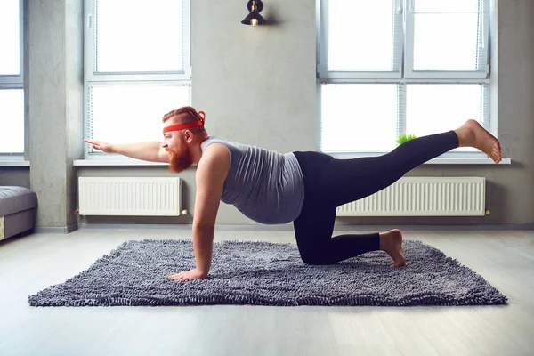 Een grappige vet bebaarde man in sport kleding doet yoga op de kamer. — Stockfoto