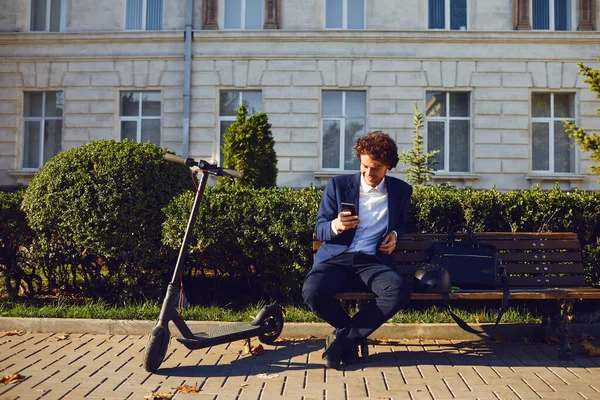 A young curly man sits on a bench with an electric scooter