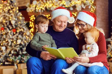 Grandparents reading a book with a child in the room with tree on Christmas Day. clipart